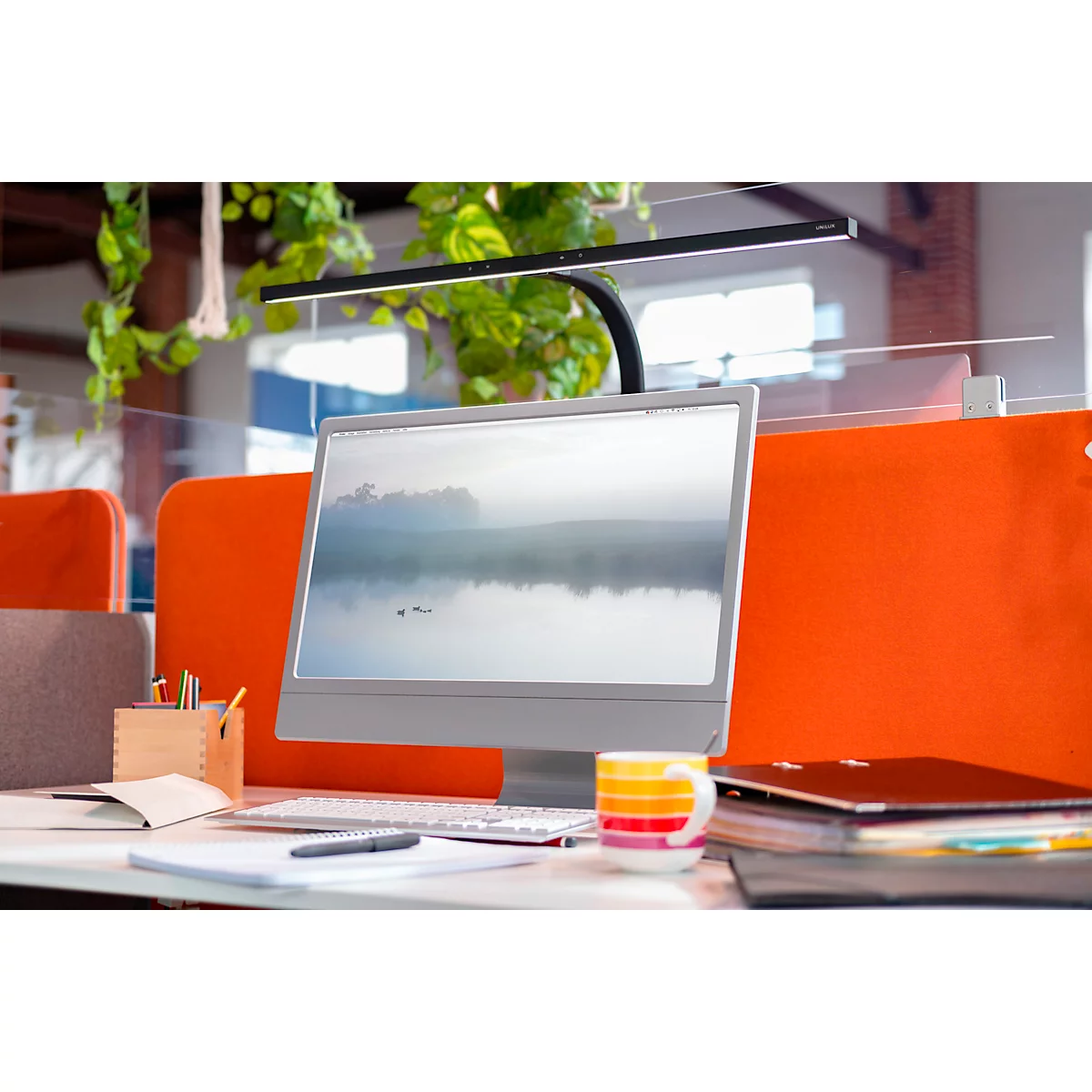 Schreibtisch mit Bildschirm, Tastatur, Tasse und Dokumenten. Darüber hängt eine Lampe. Im Hintergrund ist Grünpflanze.