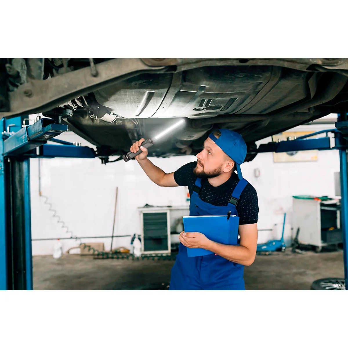 Mechanic in blue overalls inspects the undercarriage of a car. He holds a flashlight and a folder.