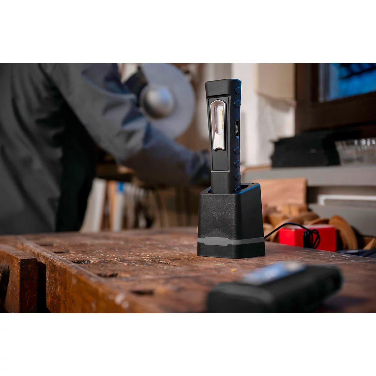 Black LED work light in charging station on wooden surface. In the background, a person is working with tools.