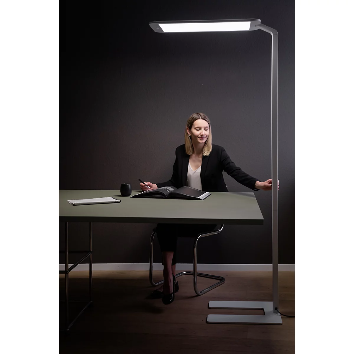 Une femme est assise à un bureau sous une lampe sur pied, regardant un livre ouvert. Elle porte une veste sombre.