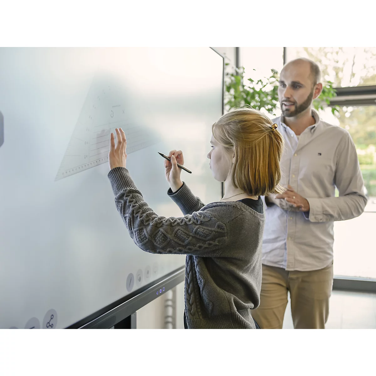 Une femme dessine avec un stylo sur un grand tableau blanc. Un homme regarde.