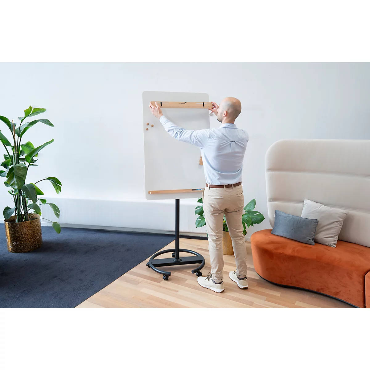 Un homme fixe un tableau blanc. Il se tient devant un fond blanc, d'autres meubles de bureau sont visibles.