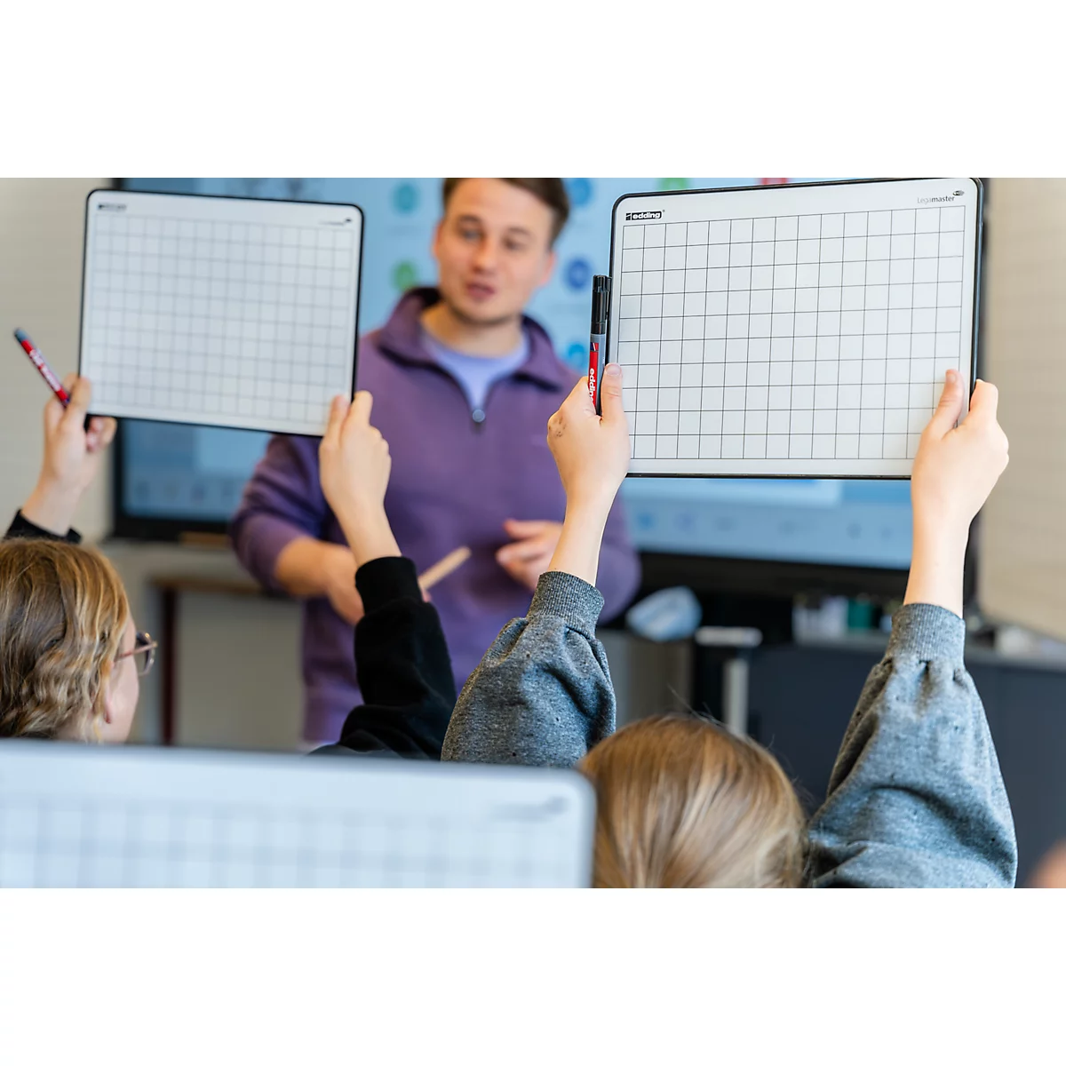 Schüler halten kleine, weiße Whiteboards mit Karomuster in die Höhe, im Hintergrund steht ein Lehrer im Klassenzimmer.
