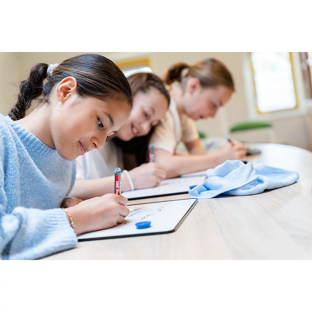 Drei Mädchen sitzen in einer Reihe an einem Tisch und schreiben konzentriert mit Stiften auf kleine Whiteboards.