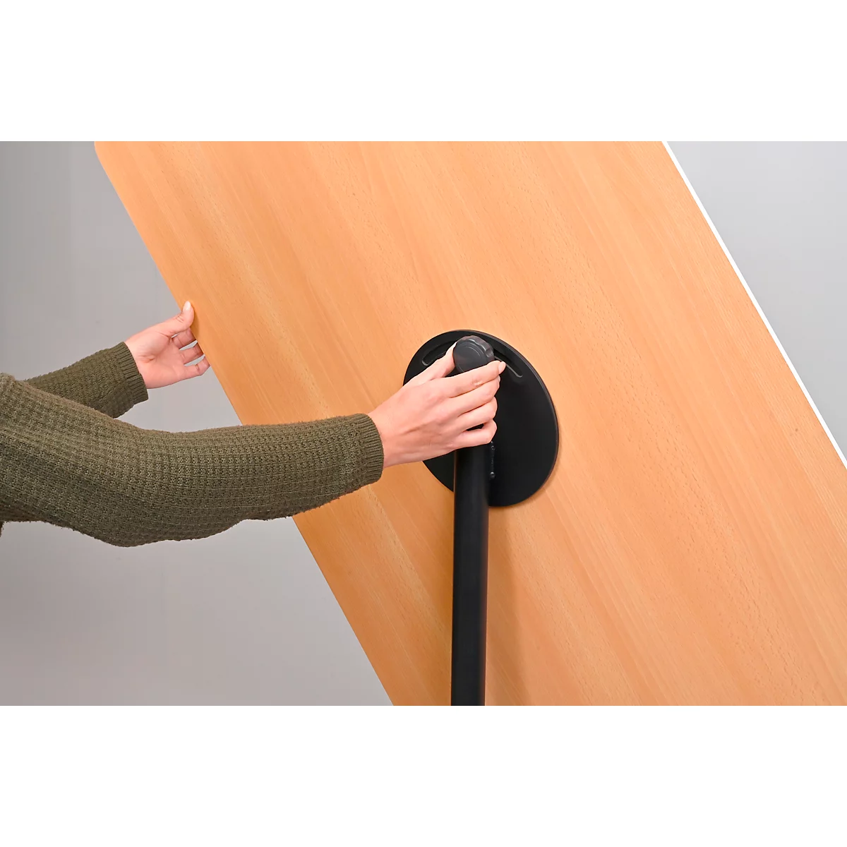 Een persoon houdt het tafelblad van een tafel vast om het op te vouwen. De tafel heeft een ronde zwarte voet en een licht houten blad.