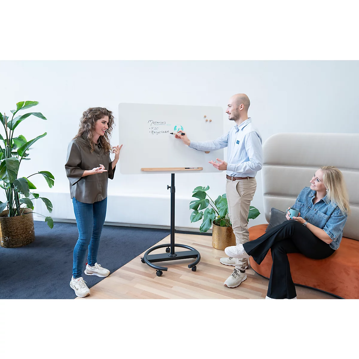 Drei Personen bei einer Präsentation. Eine Frau deutet auf das Whiteboard, ein Mann schreibt, eine dritte Person sitzt entspannt.