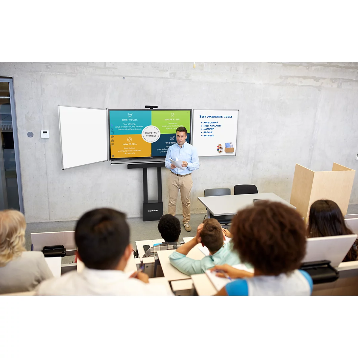 Ein Mann präsentiert vor Studenten; im Hintergrund eine Präsentation auf verschiedenen Bildschirmen.