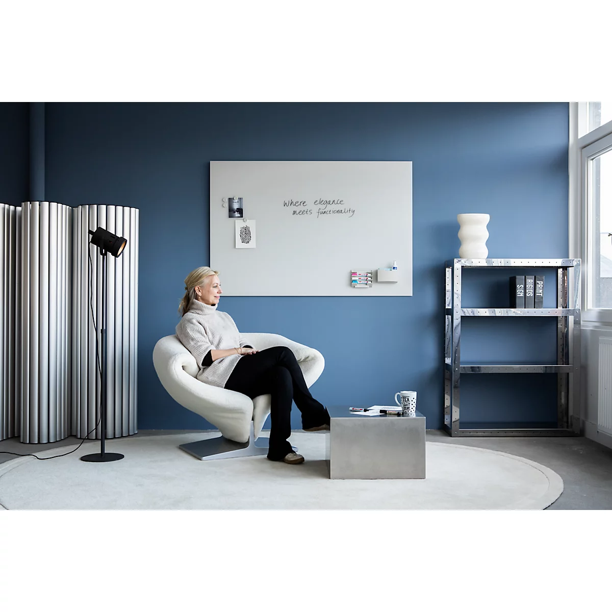 Une femme est assise dans un fauteuil blanc dans une pièce bleue. Un tableau blanc affiche "Where ideas meet ingenuity".