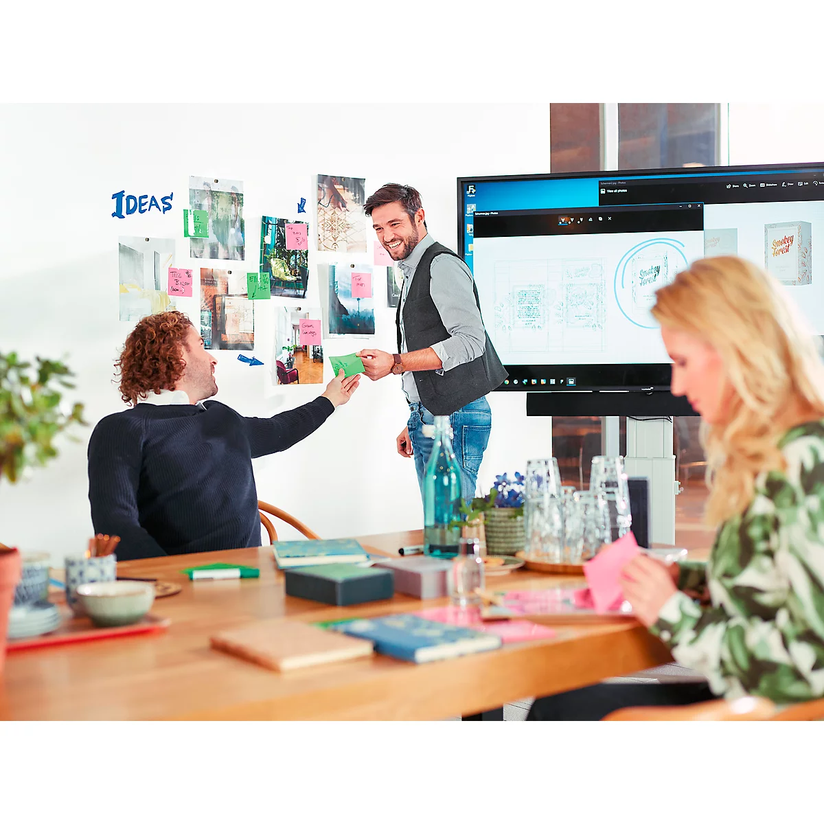 Three people at a presentation in the office. Two men exchange notes, a woman works at the table.
