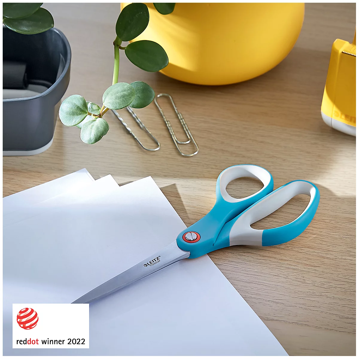 Turquoise en witte schaar die papier snijdt op een houten tafel. Daarnaast: planten, paperclips en een rood Red Dot Winner logo.
