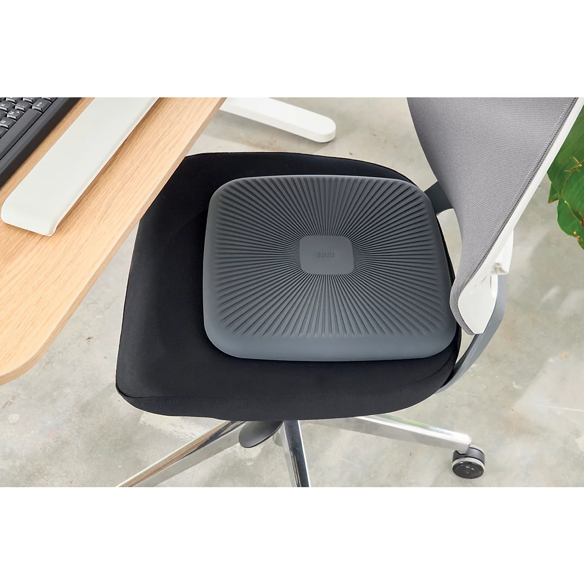 Un coussin de siège carré gris avec des rainures radiales est posé sur une chaise de bureau noire. À droite du dossier de la chaise se trouve un mur blanc.