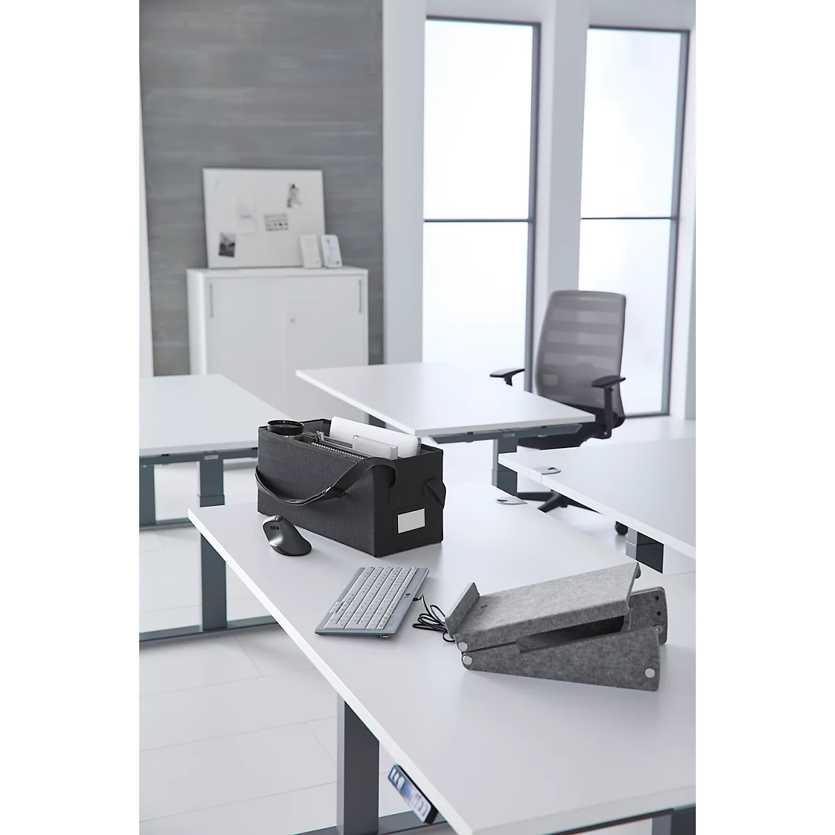 Een bureau met accessoires, zoals een toetsenbord, muis en tas. Op de achtergrond ramen en een bureaustoel. Lichte, moderne sfeer.