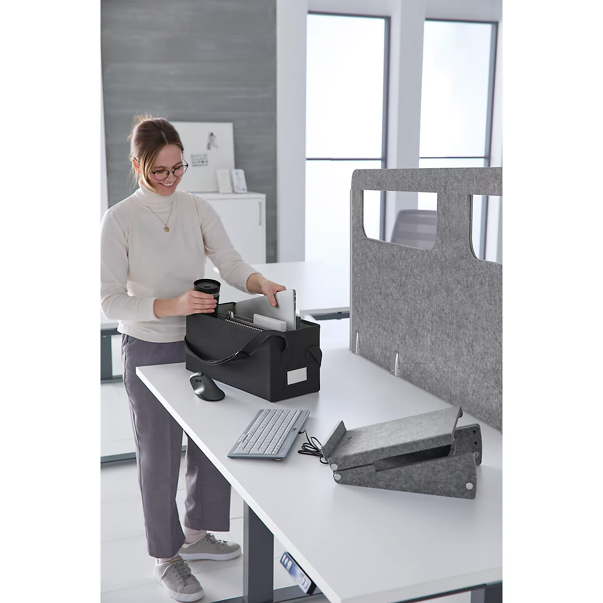 Frau am Schreibtisch mit schwarzer Tasche. Sie legt Gegenstände hinein. Auf dem Tisch liegen Maus, Tastatur und graue Unterlagen.