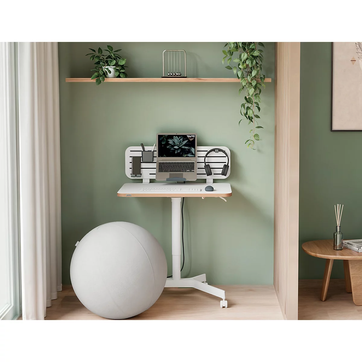 Desk with laptop, headphones, and pens. Next to it, an exercise ball, in the background a green wall and shelf with plants.