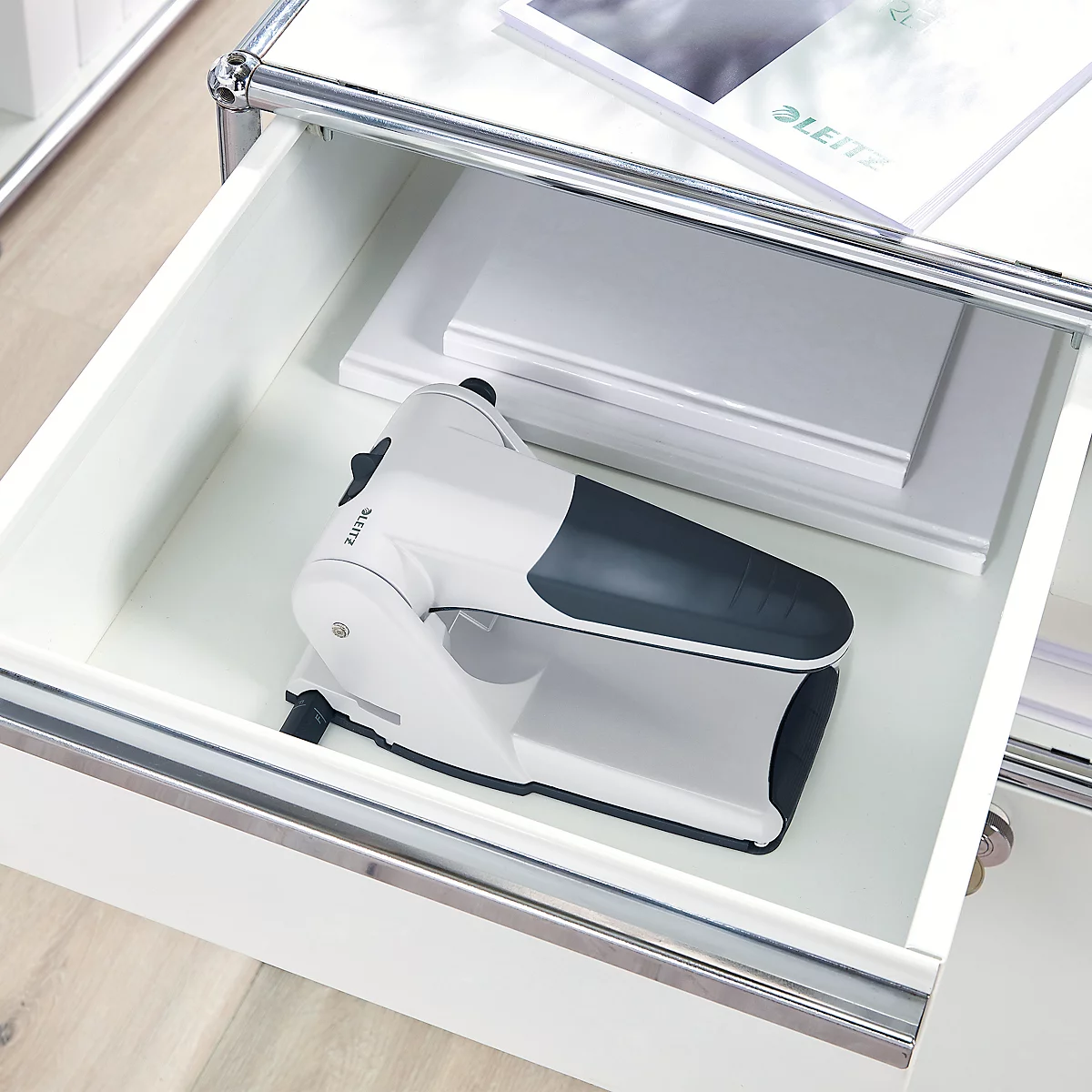 A white hole punch with a dark tip in a drawer. Papers and brochure next to it.