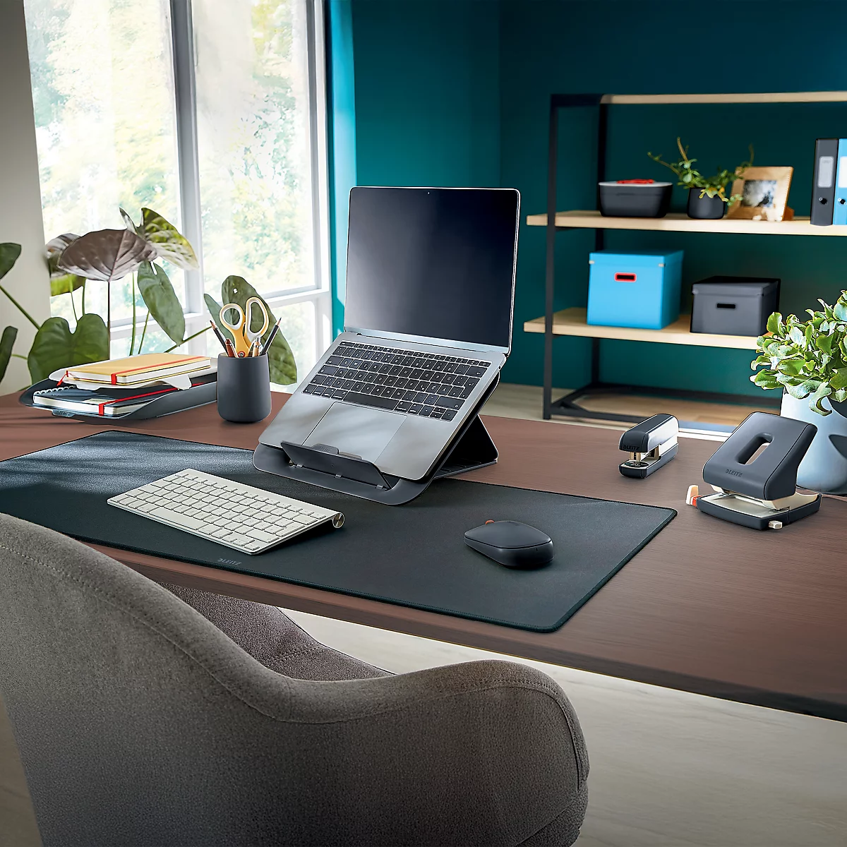 Bureau met laptop, muis, toetsenbord en perforator; raam en plank in de achtergrond.