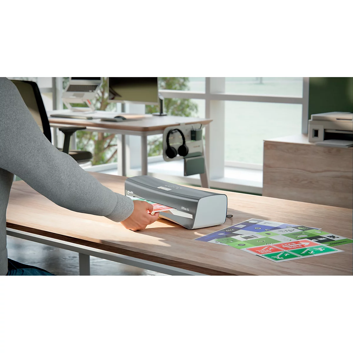 Eine Person legt ein Dokument in ein Laminiergerät auf einem Schreibtisch. Im Hintergrund: Fenster, Monitor, Kopfhörer.