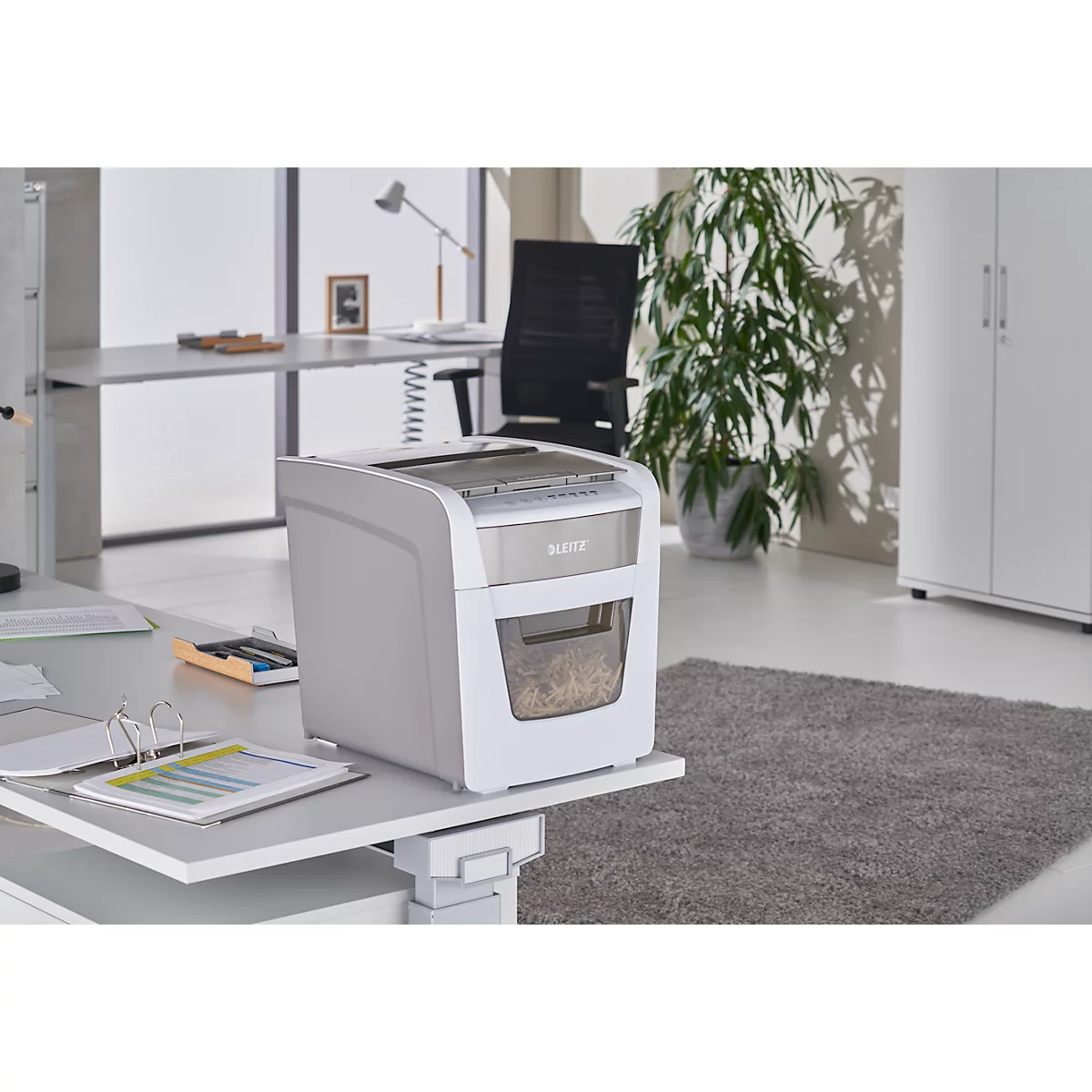 White paper shredder on a desk. In the background an office with plant, desk and cabinet.