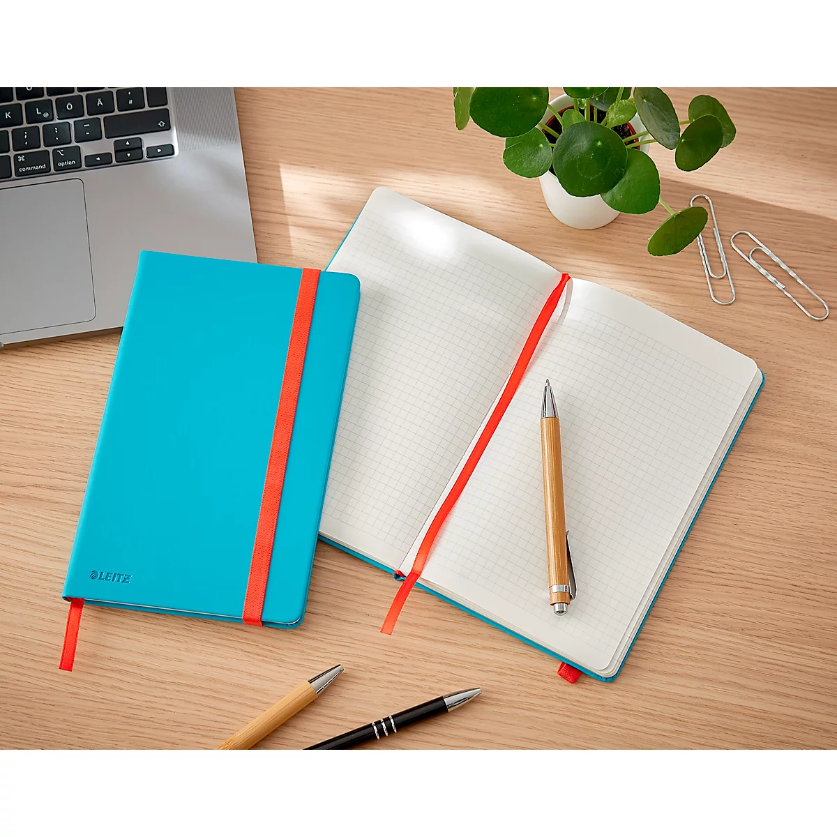 Turquoise notitieboek met rode band naast een open wit geruit notitieboek met rode bladwijzer, pen.
