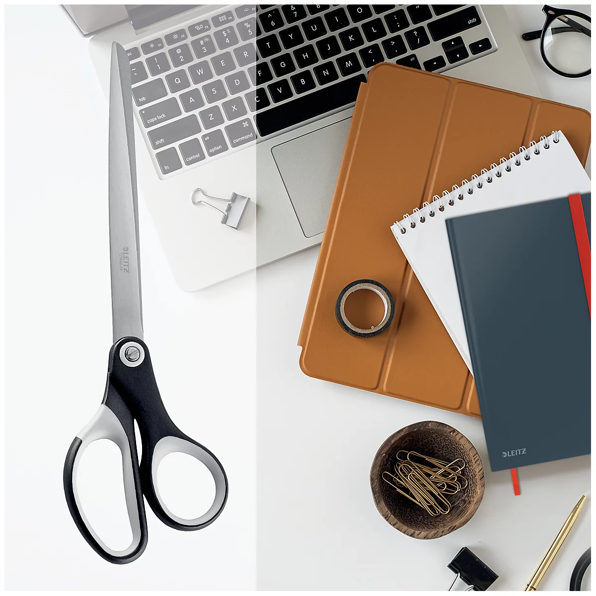 Große Schere auf einem weißen Schreibtisch, daneben ein Laptop, Notizbücher und Büroaccessoires.