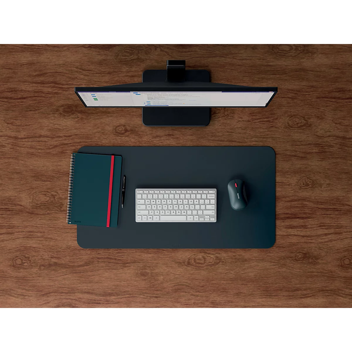 Draufsicht auf einen Schreibtisch mit Monitor, Maus, Tastatur, Notizbuch und Stift. Die Elemente liegen auf einer dunklen Schreibtischunterlage.