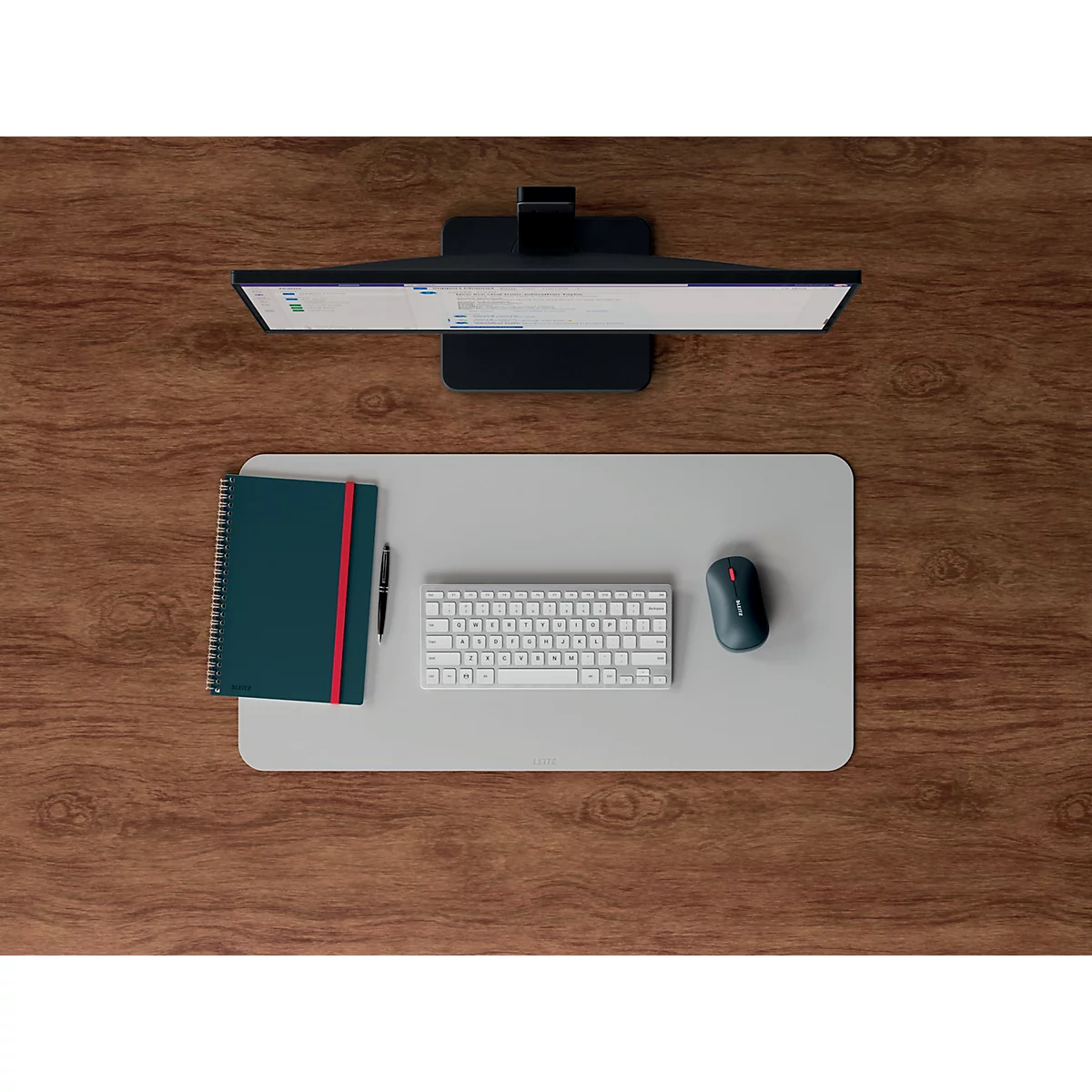 Vue de dessus d'un bureau. Moniteur, souris, clavier, cahier et stylo sont sur un tapis gris clair sur une surface en bois brun.