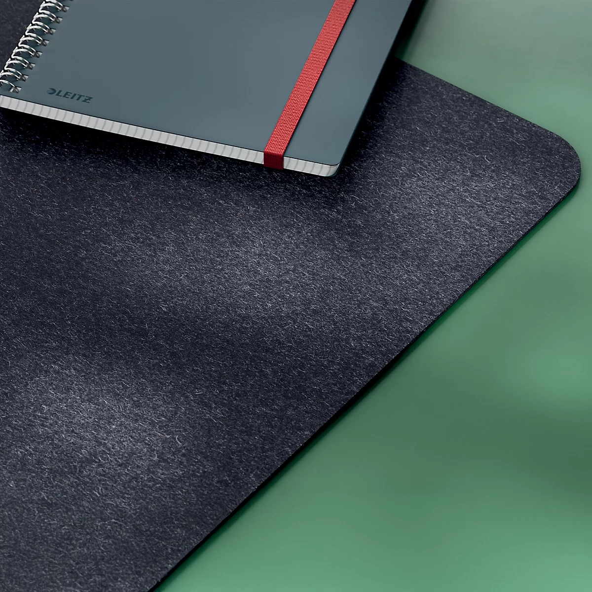 Cahier gris foncé sur surface noire, élastique rouge. Sur fond vert.