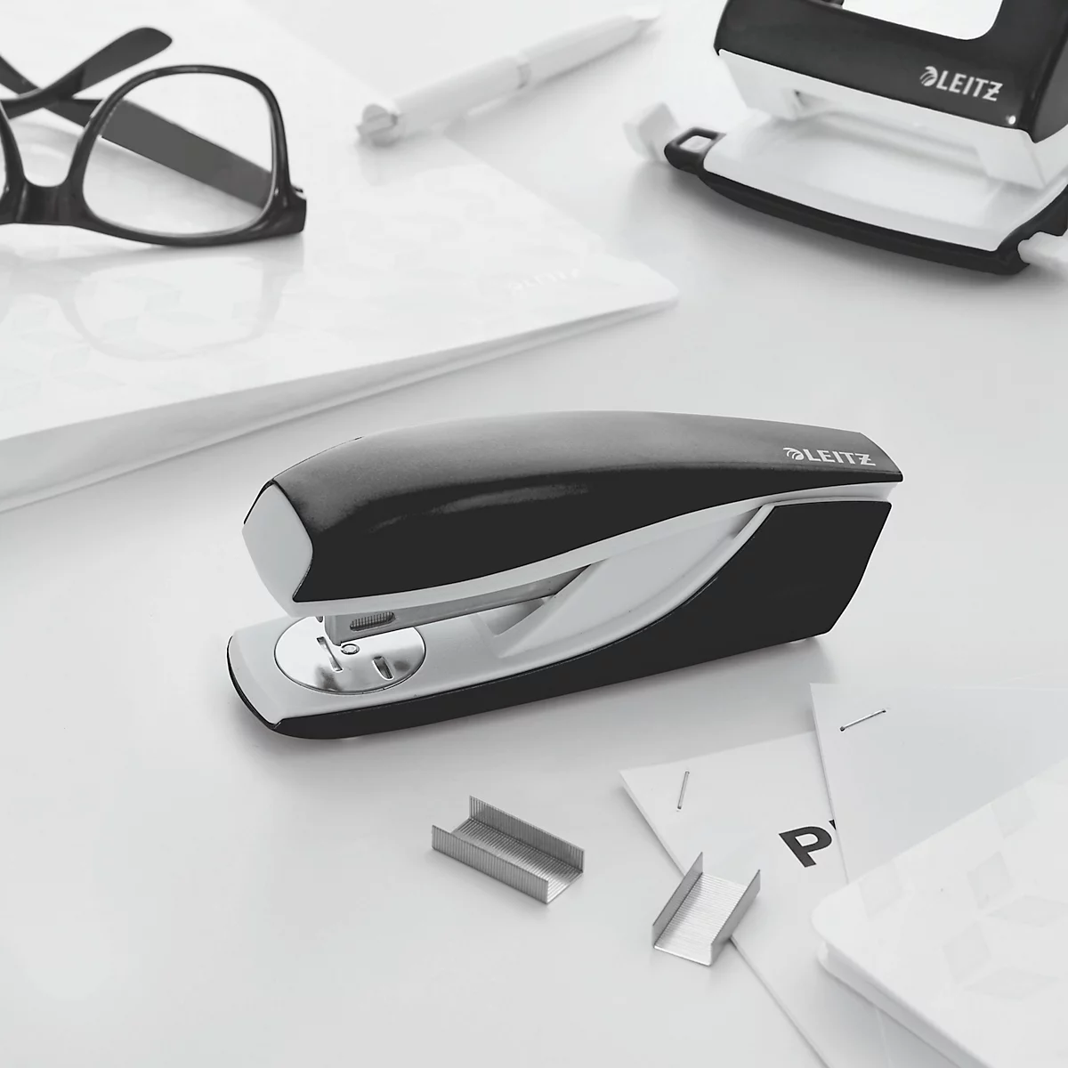 Black stapler with white accents, pens, glasses, hole punch, and paper on a white table. LEITZ logo.