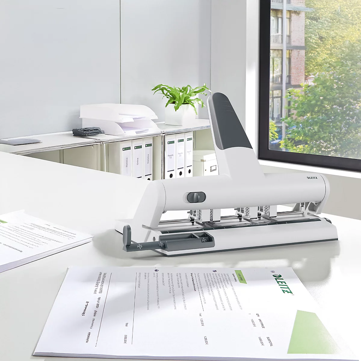 White hole punch on a desk with paper. In the background office furniture and a window with a view of greenery.