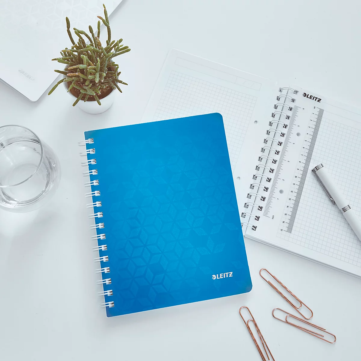 Blauw notitieboek op een witte tafel. Daarnaast een liniaal, pen, paperclips en een potplant. Op de cover: 'LEITZ'.