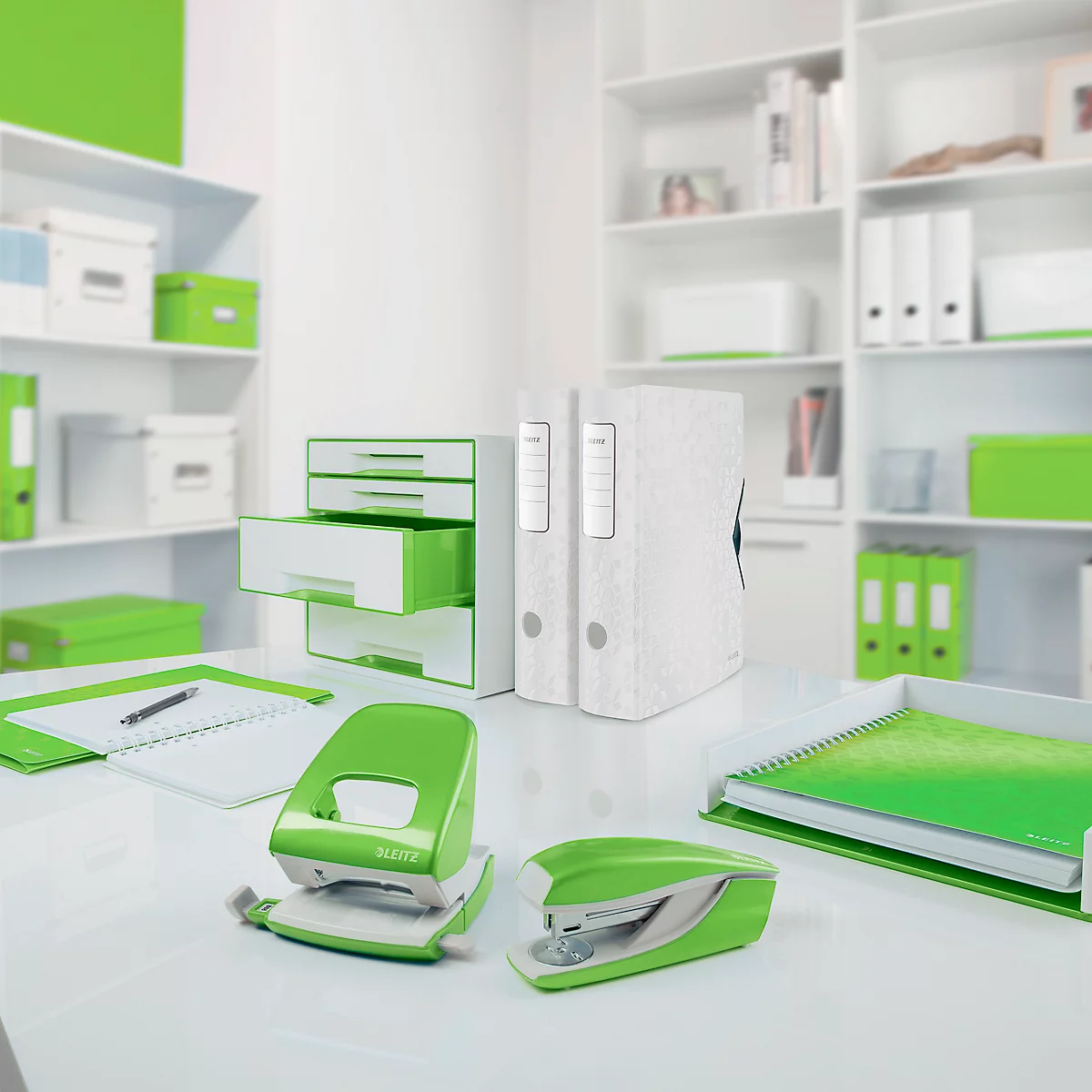 Kantooropstelling met een witte bureau, groene accenten en kantoorbenodigdheden zoals een perforator, nietmachine, ordners en notitieboek.
