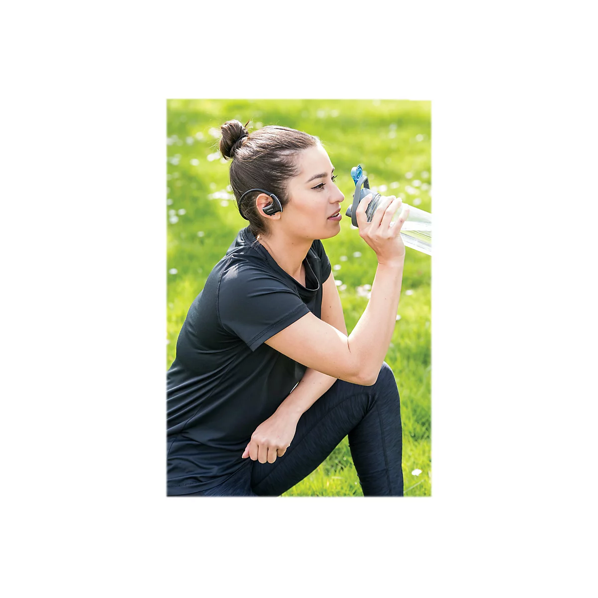 Eine Frau trinkt Wasser aus einer Flasche, sie trÃ¤gt KopfhÃ¶rer und sitzt im Gras.