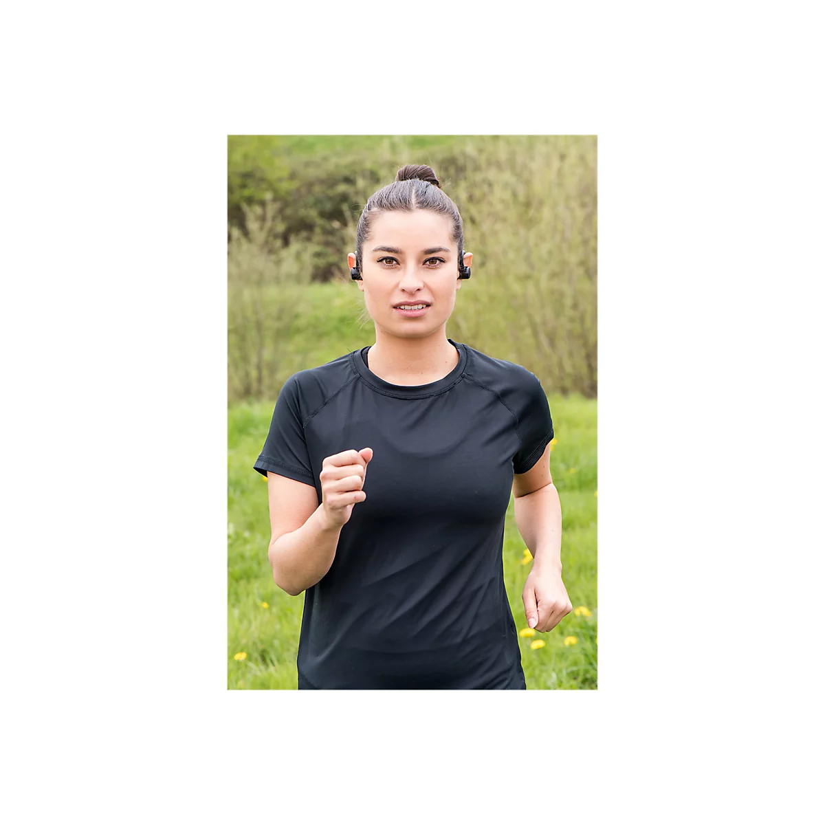 LÃ¤uferin mit KopfhÃ¶rern im schwarzen Shirt. Sie blickt in die Kamera, im Hintergrund Wiese und BÃ¤ume.