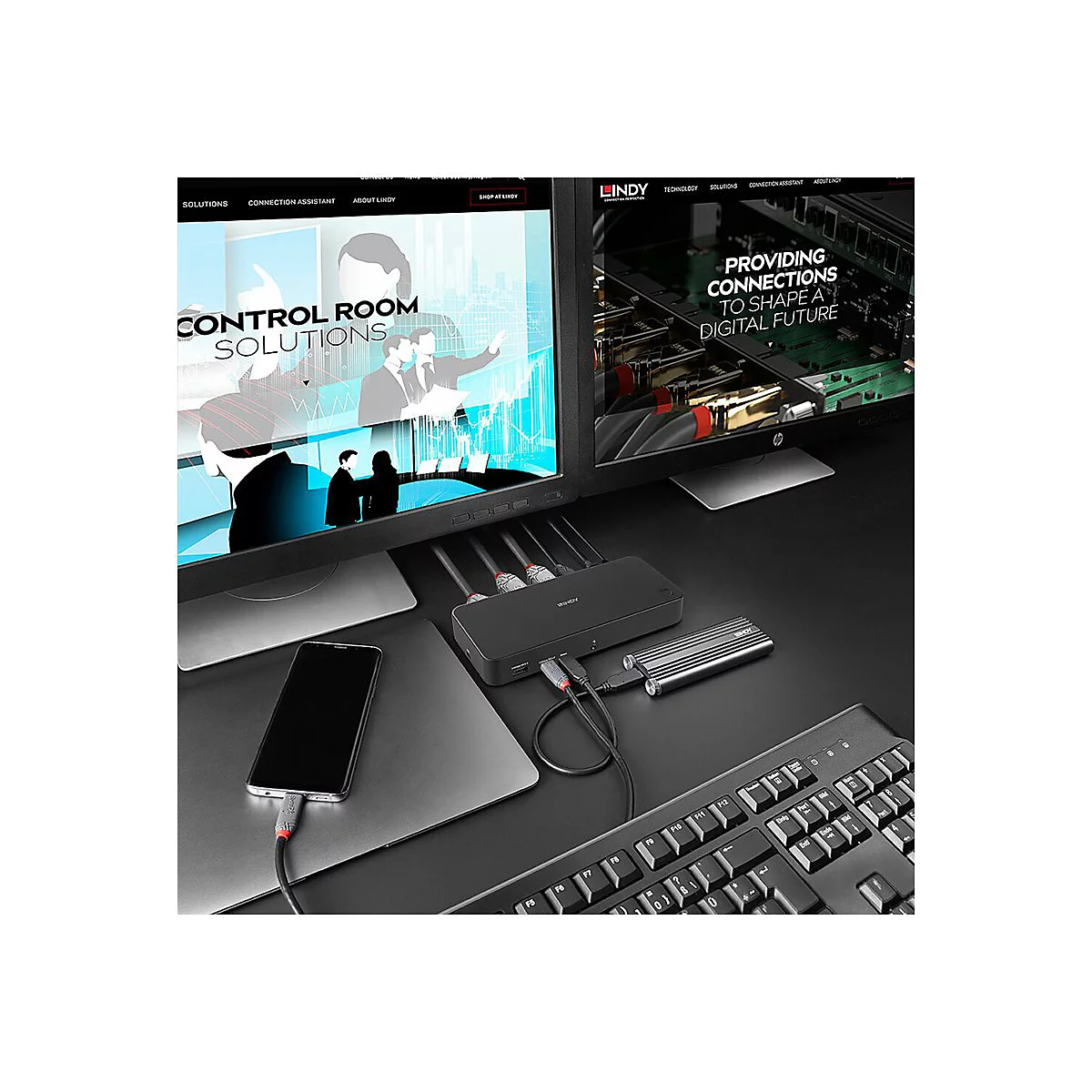 Schwarzer Hub mit Kabeln, Laptop und Smartphone auf einem Schreibtisch. Zwei Monitore zeigen Webseiten. Tastatur im Vordergrund.