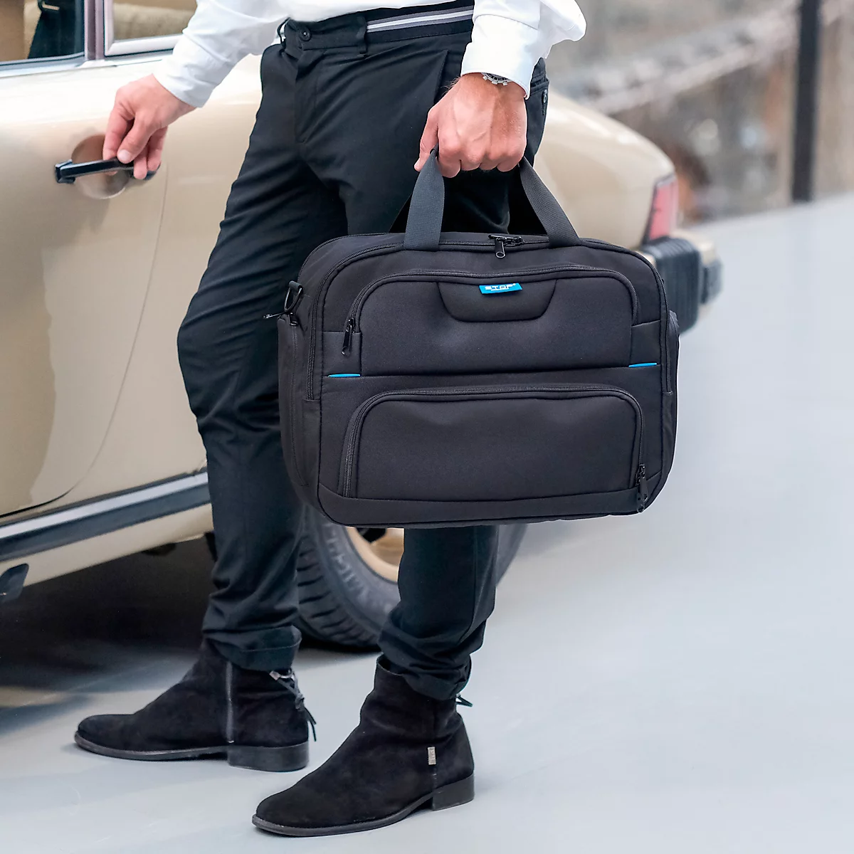 Un homme en pantalon et bottes noirs tenant une mallette noire. Il ouvre la portière d'une voiture ancienne de couleur claire.