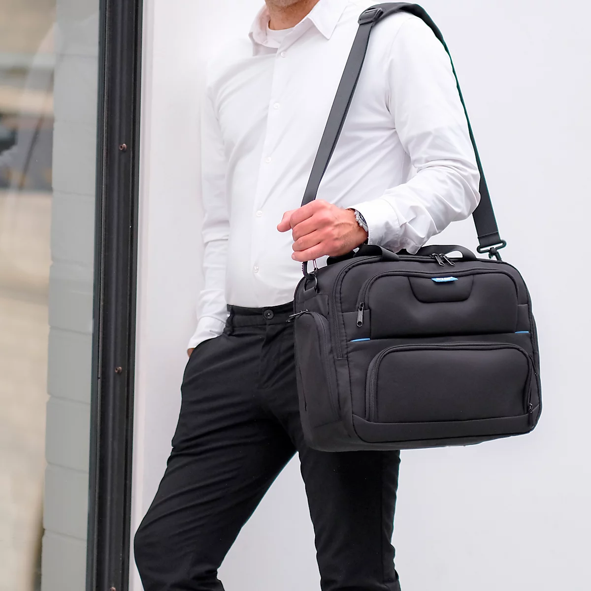 Un homme portant une mallette noire avec une bandoulière, une chemise blanche et un pantalon noir. Appuyé contre un mur blanc.