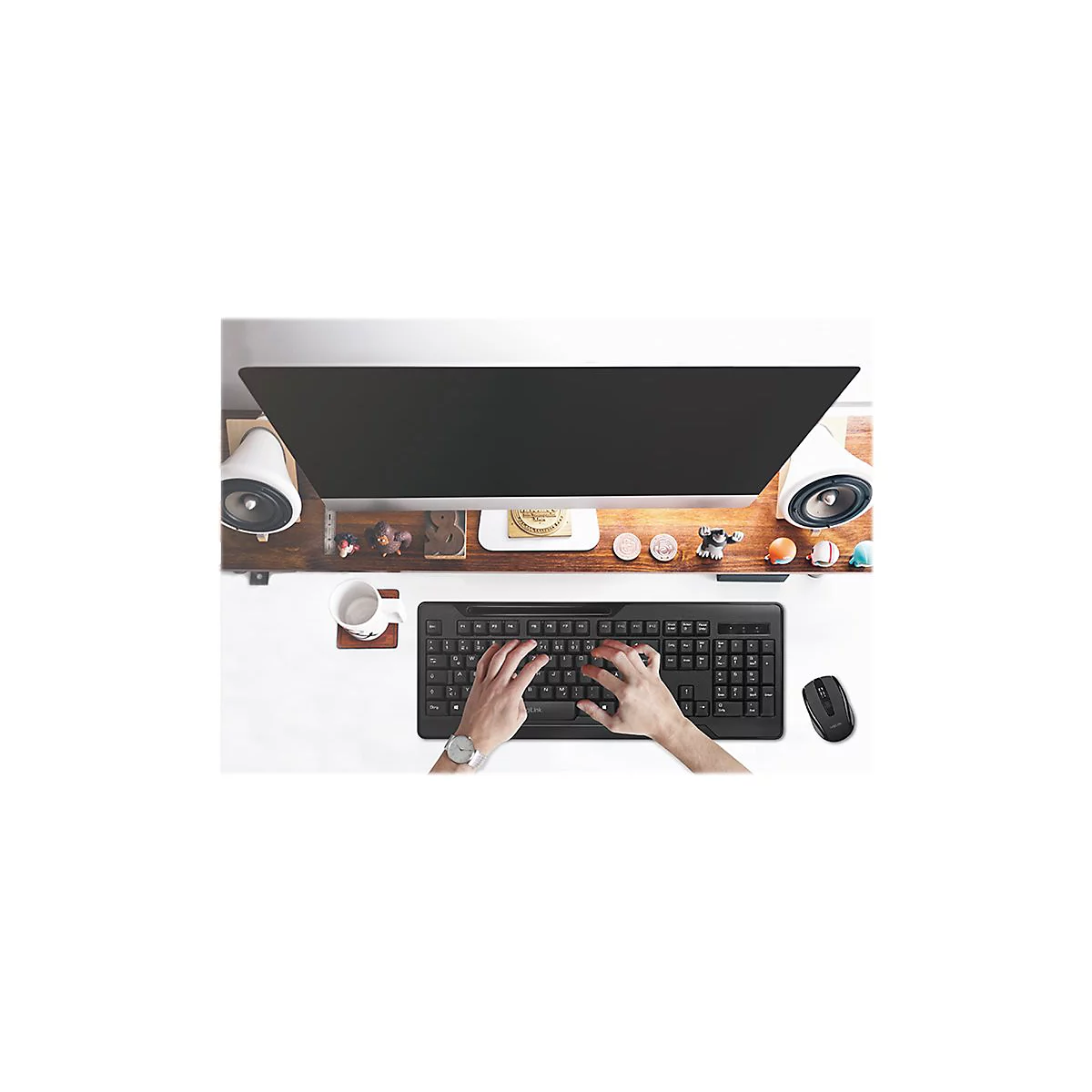 Person tippt auf schwarzer Tastatur. Auf dem Tisch: Monitor, Lautsprecher, Maus, Tasse, Deko.
