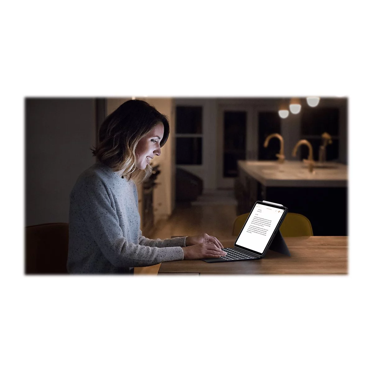 Eine Frau arbeitet abends am Laptop. Sie sitzt an einem Holztisch, das Tablet ist beleuchtet. Im Hintergrund ist eine KÃ¼che zu sehen.
