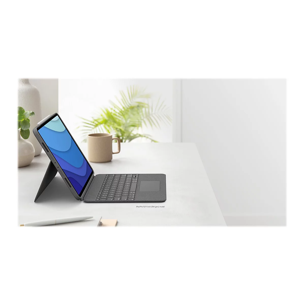 Ein Tablet mit Tastatur auf einem Tisch. Der Bildschirm zeigt blaue Wellen. Rechts eine Tasse, links Pflanzen und Stifte.