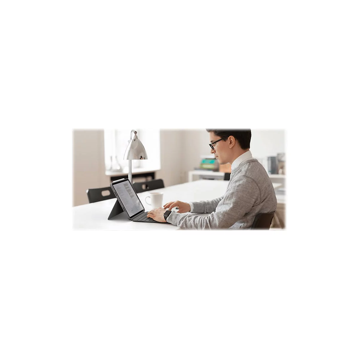 Ein Mann mit Brille tippt an einem Tablet mit Tastatur, während er an einem Tisch sitzt. Eine Lampe, eine Tasse und ein Fenster sind im Hintergrund zu sehen.