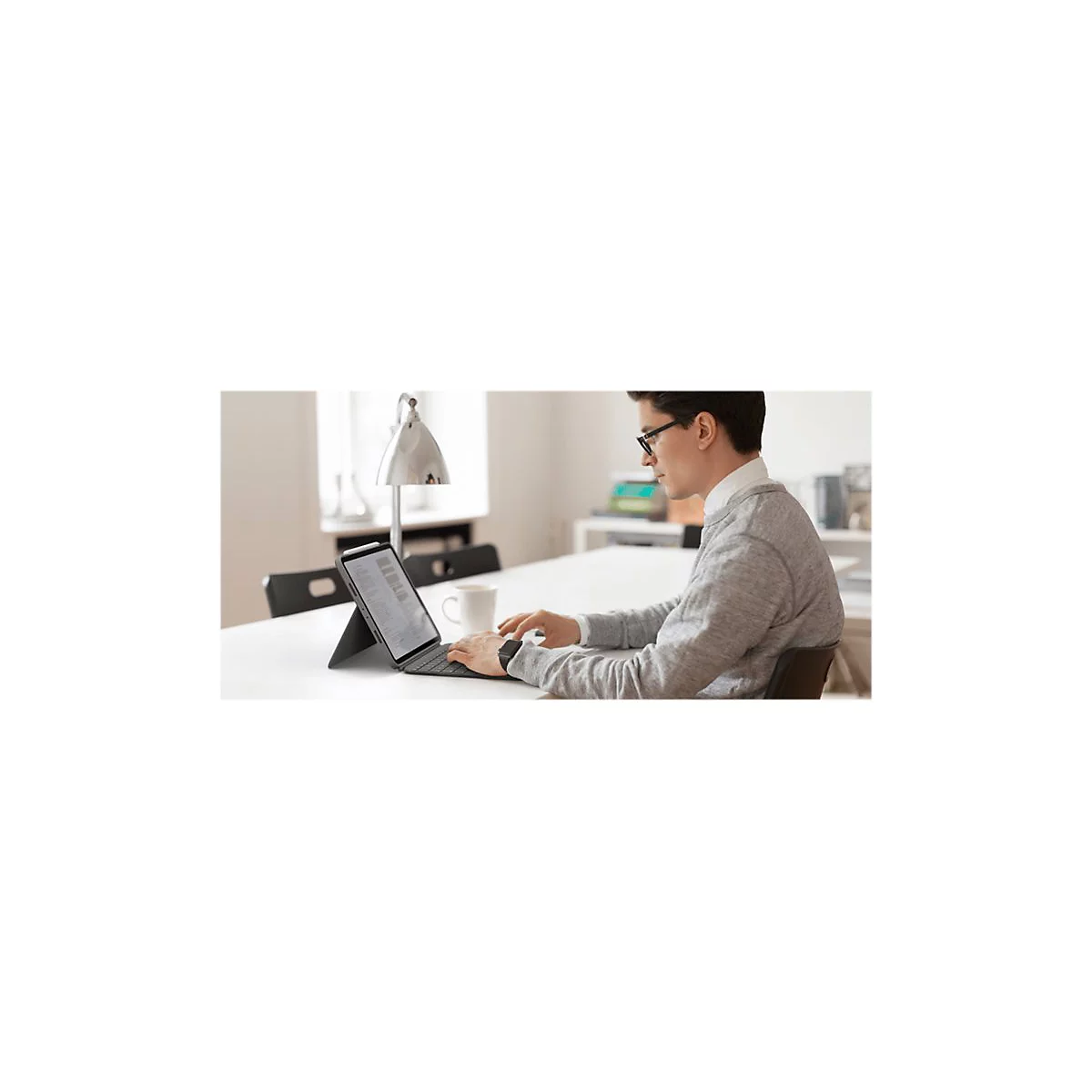 Mann sitzt an Tisch, tippt auf Tablet mit Tastatur. Er trägt Brille, grauen Pullover und weißes Hemd. Auf dem Tisch steht eine Tasse.