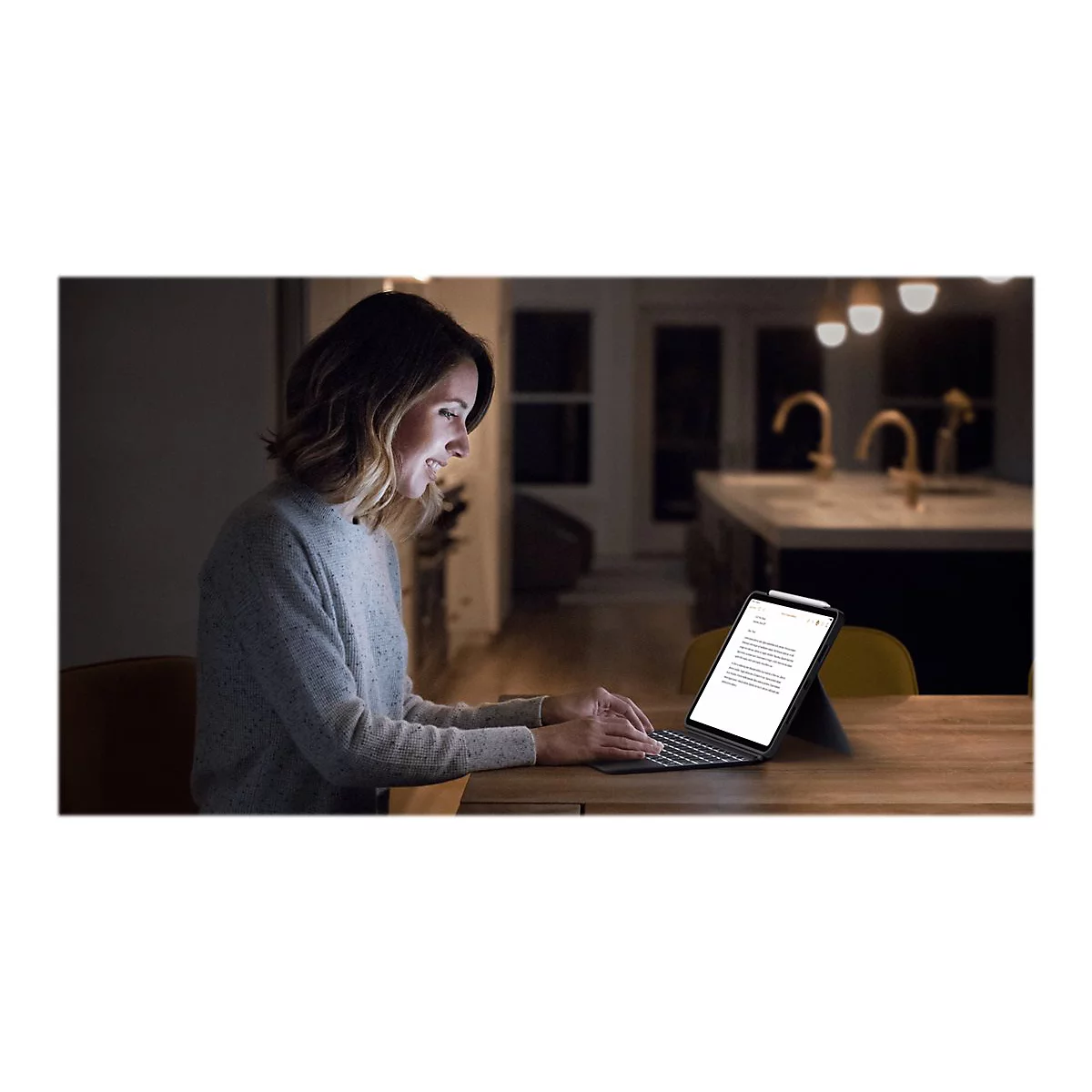 Eine Frau tippt abends an einem Tablet mit angeschlossener Tastatur. Die Frau lächelt und blickt auf das Tablet. Im Hintergrund ist eine Küche zu sehen.