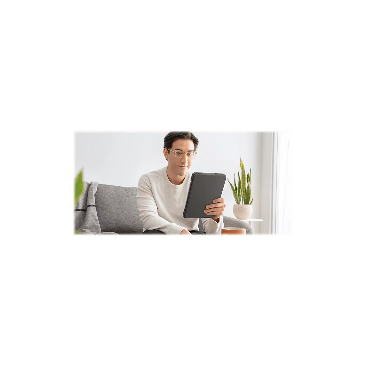 Ein Mann mit Brille sitzt auf einem Sofa und betrachtet ein Tablet. Neben ihm steht eine Topfpflanze.