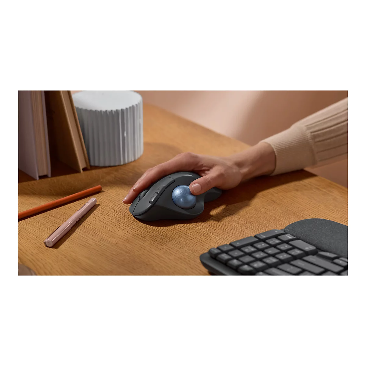 Dunkelgraue ergonomische Maus mit blauer Kugel in einer Hand. Auf Holzoberfläche. Eine Tastatur und Stifte liegen daneben.