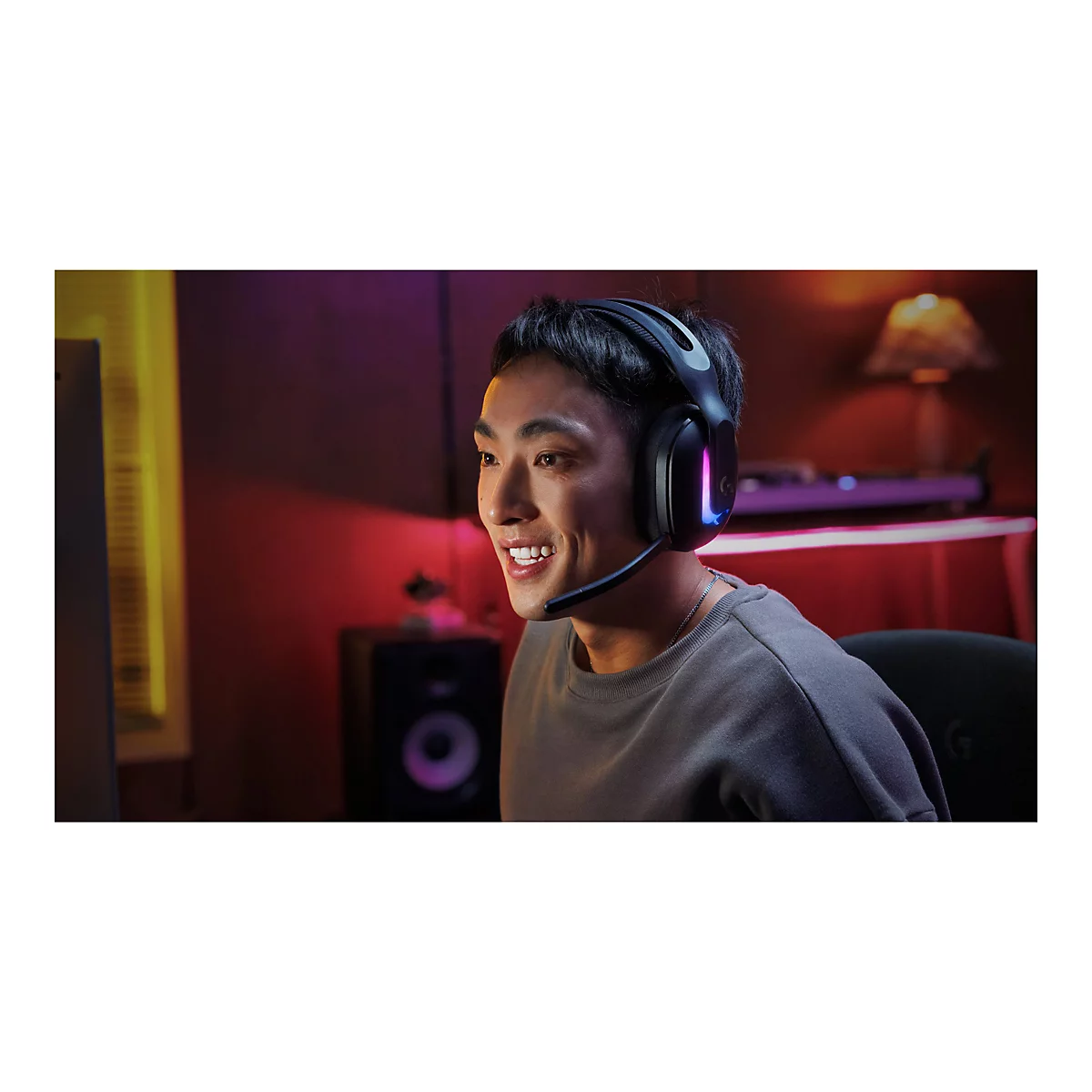 Ein junger Mann mit Headset, lächelnd vor einem Bildschirm. Das Headset ist schwarz mit farbigen Lichtern, er sitzt in einem Gaming-Setup.
