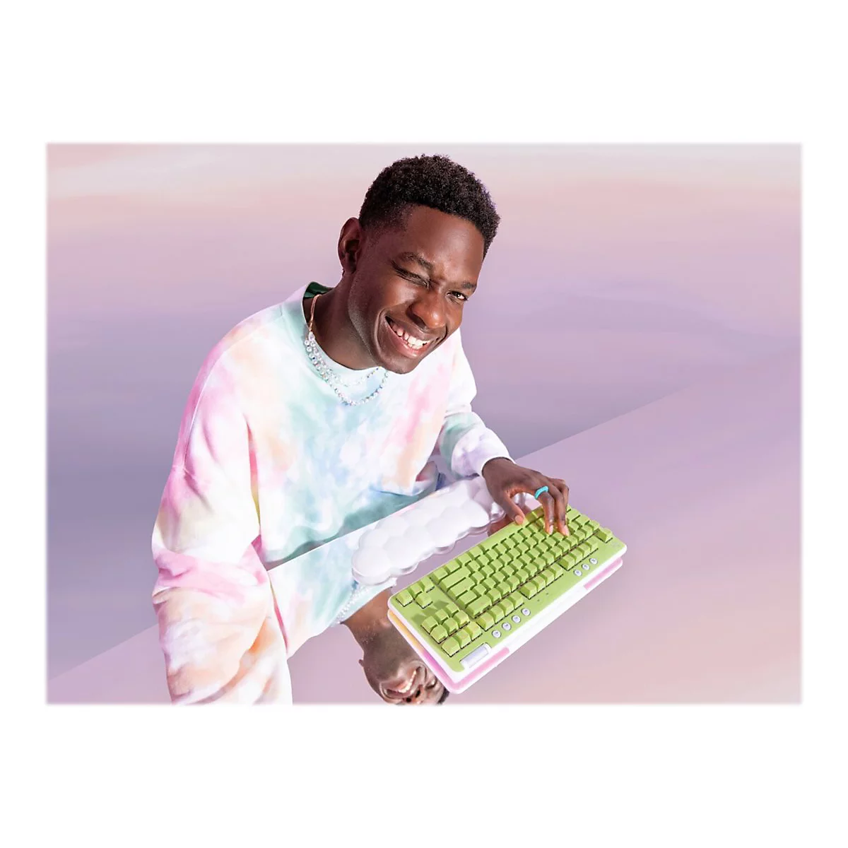Ein Mann mit Regenbogen-Sweatshirt hält grüne Tastatur; er lächelt und zwinkert. Hintergrund ist zweifarbig.