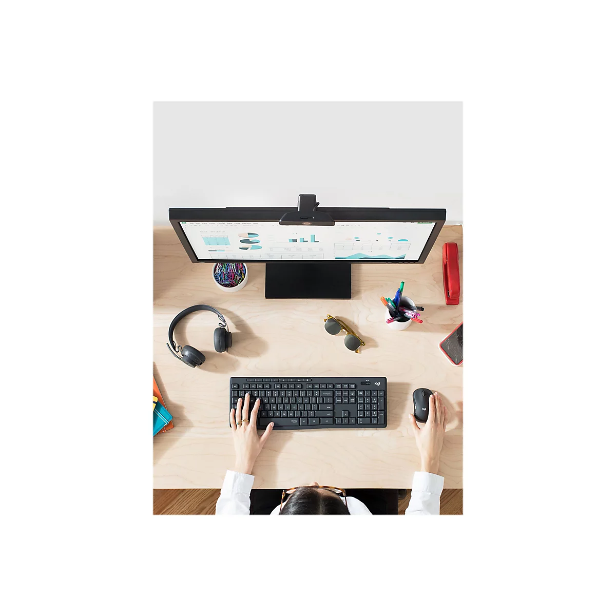 Draufsicht auf einen Schreibtisch mit Monitor, Tastatur, Maus, Kopfhörern, Brille und Stiften. Eine Person tippt auf der Tastatur.
