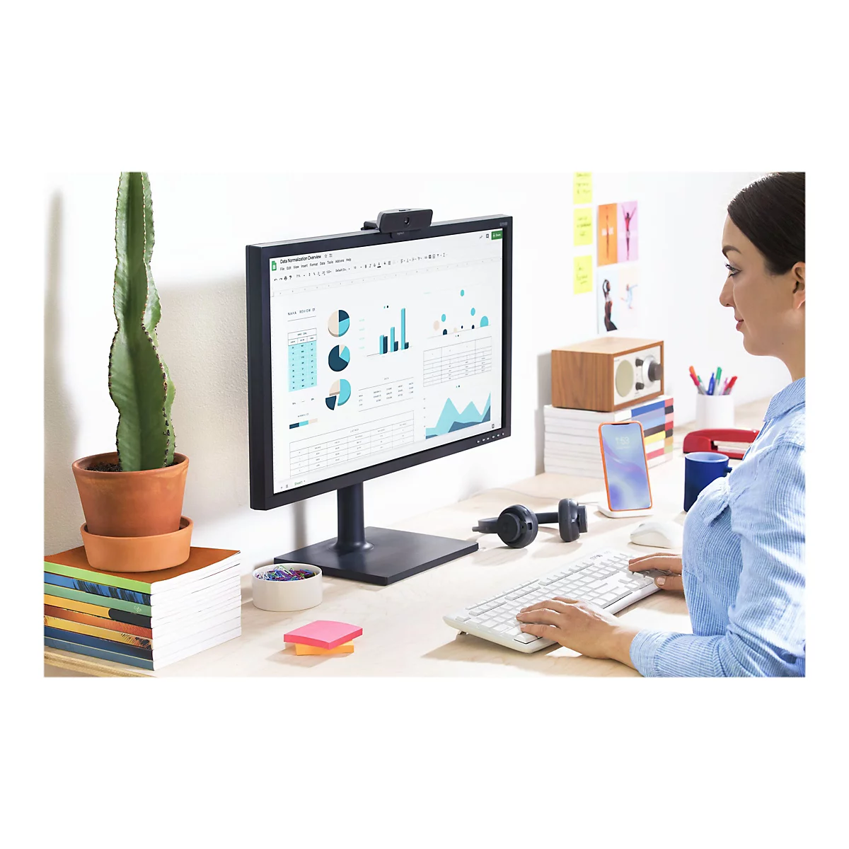 Eine Frau sitzt an einem Schreibtisch und arbeitet mit einem Monitor. Auf dem Bildschirm sind Diagramme zu sehen. Auf dem Tisch ein Kaktus, Smartphone.