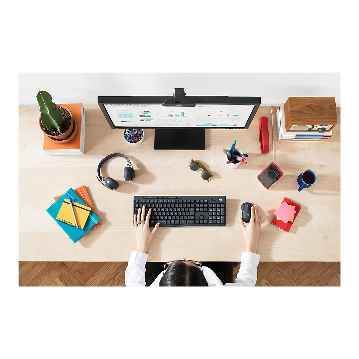 Draufsicht auf einen Schreibtisch: Monitor, Tastatur, Maus, Kopfhörer, Sonnenbrille, Notizblöcke, Stifte, Kaktus, Handy, Tasse.