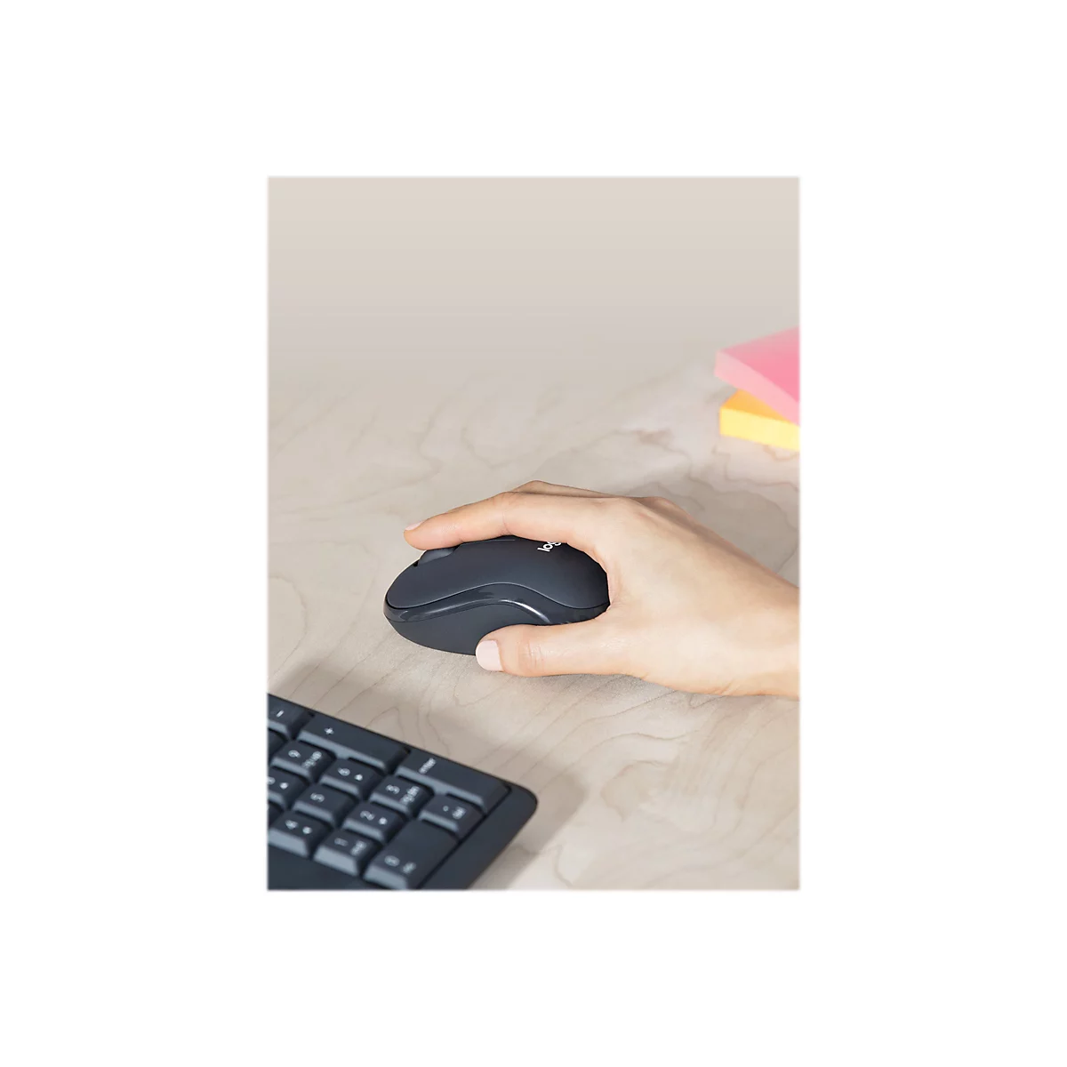Eine Hand liegt auf einer schwarzen Maus, daneben ein schwarzes Keyboard auf einem Holzschreibtisch. Im Hintergrund liegen gelbe und pinkfarbene Haftnotizen.